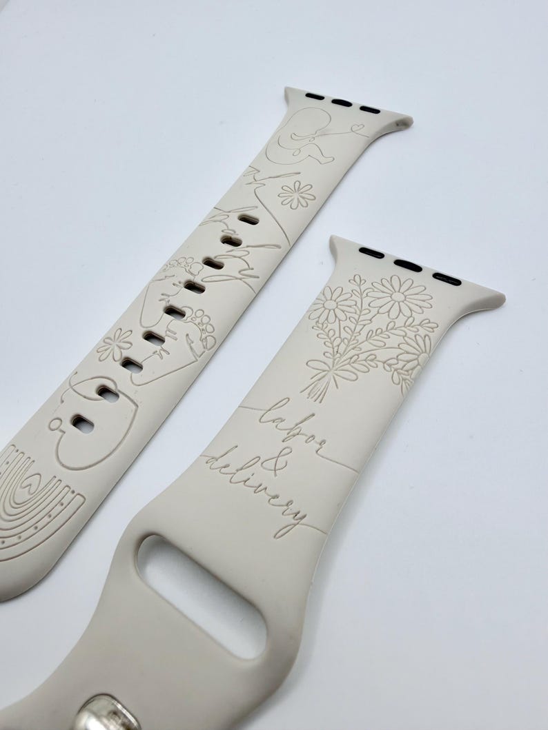 May include: A light beige silicone watch band featuring engraved line art designs. One strap displays a fetus, flowers, and a rainbow. The other strap features a bouquet of daisies and the cursive text labor & delivery, with black metal connectors.