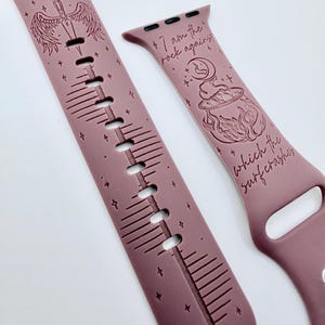 May include: A mauve silicone watch band featuring engraved designs. One side displays a winged sword with the text "VALKYRIE nothing can BREAK ME" and star accents. The other side shows a cauldron and moon with the text "I am the rock against which the surf crashes".