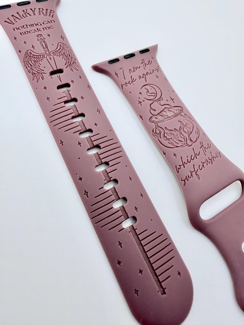 May include: A mauve silicone watch band featuring engraved designs. One side displays a winged sword with the text "VALKYRIE nothing can BREAK ME" and star accents. The other side shows a cauldron and moon with the text "I am the rock against which the surf crashes".