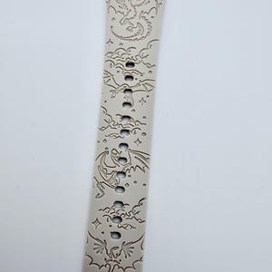 May include: A light beige silicone watch band featuring an engraved pattern of multiple dragons flying among clouds and stars. The strap includes a series of vertical adjustment holes running down the center, with a curved connector end piece.