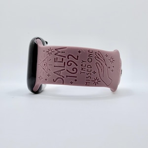 May include: A dusty rose silicone watch band featuring an embossed design with the text SALEM 1692 THEY MISSED ONE, accompanied by an eye in a triangle, a hand, and various star symbols, attached to a black watch casing on the left side.
