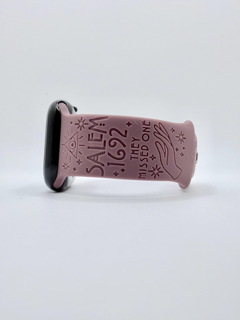 May include: A dusty rose silicone watch band featuring an embossed design with the text SALEM 1692 THEY MISSED ONE, accompanied by an eye in a triangle, a hand, and various star symbols, attached to a black watch casing on the left side.