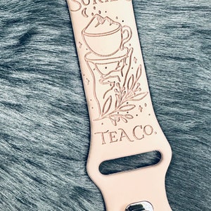 May include: A light beige silicone watch band featuring a laser-engraved design of a teacup with a mountain peak, a sprig of leaves, and the text SURIEL TEA CO. The band includes a silver-toned metal pin-and-tuck closure mechanism at the bottom.