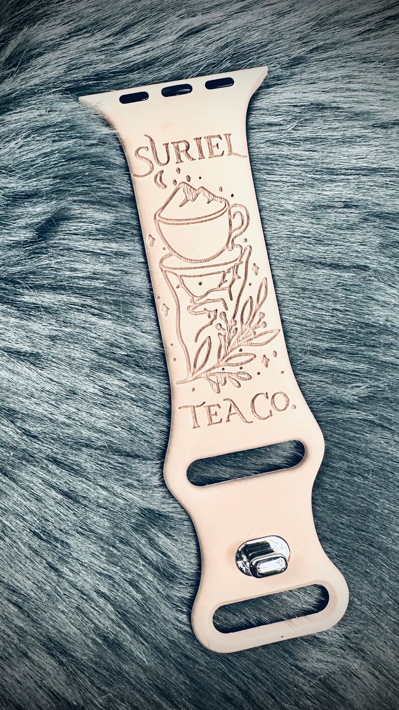 May include: A light beige silicone watch band featuring a laser-engraved design of a teacup with a mountain peak, a sprig of leaves, and the text SURIEL TEA CO. The band includes a silver-toned metal pin-and-tuck closure mechanism at the bottom.