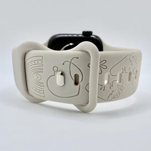 May include: A light beige silicone watch band featuring engraved line art designs including a rainbow, a stethoscope, a heart, and flowers. The band is attached to a black smartwatch case and includes a metallic silver pin closure mechanism.