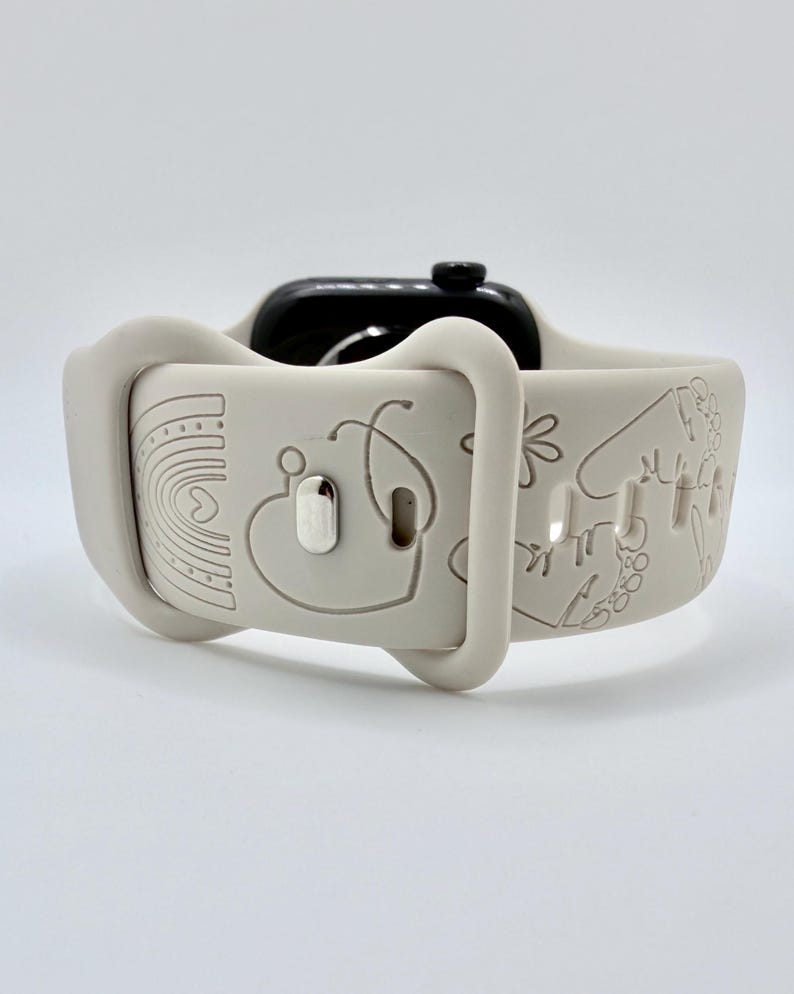 May include: A light beige silicone watch band featuring engraved line art designs including a rainbow, a stethoscope, a heart, and flowers. The band is attached to a black smartwatch case and includes a metallic silver pin closure mechanism.