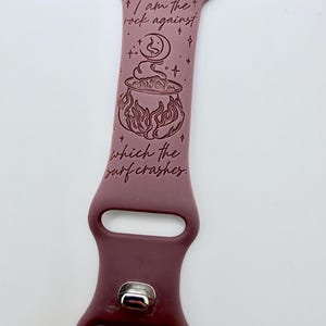 May include: A mauve silicone watch band featuring a dark engraved design of a cauldron, a crescent moon, and stars. The text reads, "I am the rock against which the surf crashes." The band includes a silver-toned metal pin closure and attachment hardware.