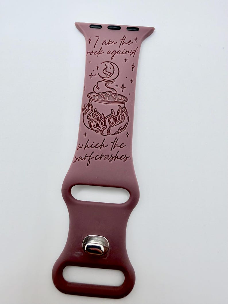 May include: A mauve silicone watch band featuring a dark engraved design of a cauldron, a crescent moon, and stars. The text reads, "I am the rock against which the surf crashes." The band includes a silver-toned metal pin closure and attachment hardware.