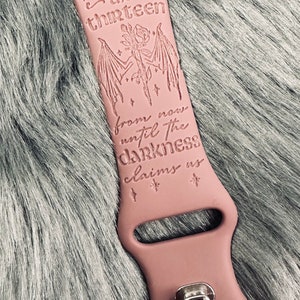 May include: A dusty rose silicone watch band featuring an engraved design of a rose with bat wings, small stars, and the text "the thirteen from now until the darkness claims us". The band includes a silver-toned metal pin closure mechanism.