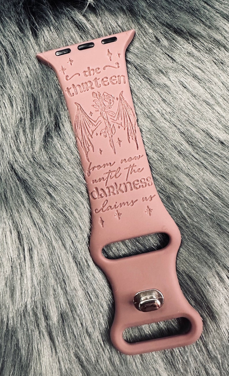 May include: A dusty rose silicone watch band featuring an engraved design of a rose with bat wings, small stars, and the text "the thirteen from now until the darkness claims us". The band includes a silver-toned metal pin closure mechanism.