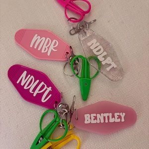 May include: A collection of keychains in various colors, including pink, green, and yellow. Some keychains feature the names "MBR", "NDLPT", and "BENTLEY" in white lettering. The keychains also include small scissor-shaped charms.