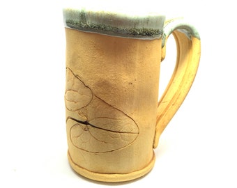 May include: A light brown ceramic mug with a green glazed rim and a handle. The mug has a raised design of leaves on the front.