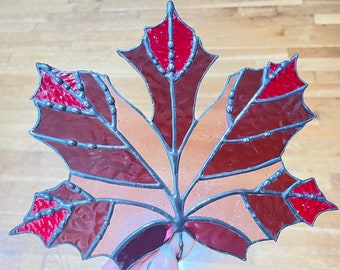May include: A stained glass maple leaf with a brown and red design. The leaf is made of glass pieces that are joined together with lead. The leaf is a decorative piece that can be hung on a wall or placed on a table.