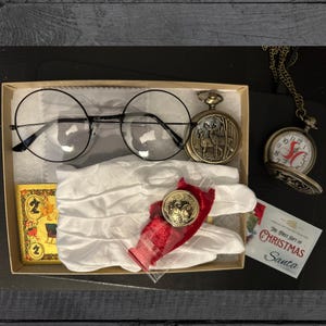 May include: A collection of vintage-style items in a box, including round-framed glasses, a pocket watch with a deer design, a small coin, a white glove, and a card that says "The First Gift of Christmas Santa."