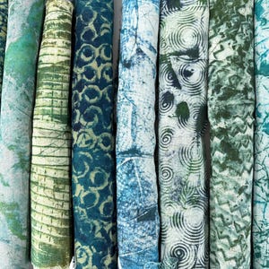 May include: A selection of rolled fabric pieces in shades of blue and green. The textiles feature a variety of patterns, such as geometric shapes, organic forms, and textured effects. The colour palette varies from light teal to deep indigo.