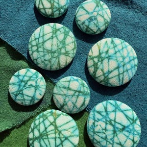May include: A collection of eight round fabric buttons with a teal and white abstract pattern. The buttons vary in size and are arranged on a teal and green fabric background. The pattern resembles a network of lines.