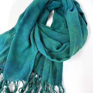 Puede incluir: Una bufanda verde y turquesa con borde de flecos. La bufanda está hecha de un material suave y tejido y tiene un efecto degradado, que pasa de un turquesa más oscuro a un verde más claro. La borla es de color turquesa a juego.