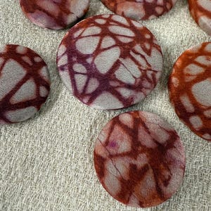 May include: Close-up of several round fabric buttons with a tie-dye pattern. The buttons feature a mix of colors, including shades of red, orange, and purple, against a light gray background. The buttons are arranged on a textured, light-colored fabric.