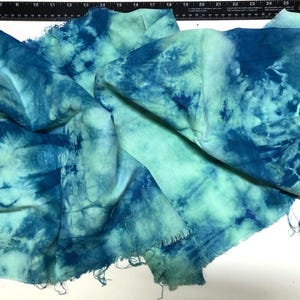 May include: A piece of fabric with a tie-dye pattern in shades of teal and blue. The fabric has a raw, unfinished edge and is draped across a surface. The dye pattern creates a marbled effect.