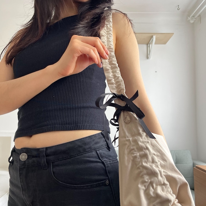 May include: A woman wearing a black tank top and black jeans is holding a beige drawstring bag with black ribbon ties.