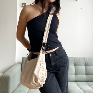 May include: A beige fabric bag with a gathered, ruffled design. The bag has a black strap with a bow detail and is worn over the shoulder of a person wearing a black one-shoulder top and black jeans.