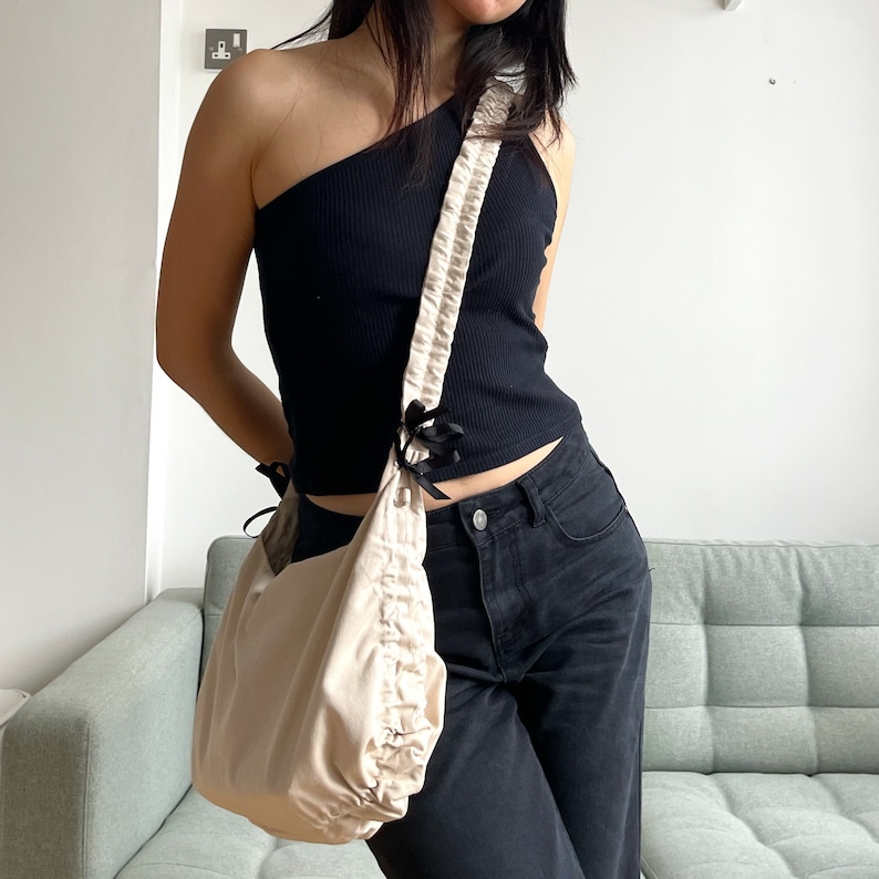 May include: A beige fabric bag with a gathered, ruffled design. The bag has a black strap with a bow detail and is worn over the shoulder of a person wearing a black one-shoulder top and black jeans.
