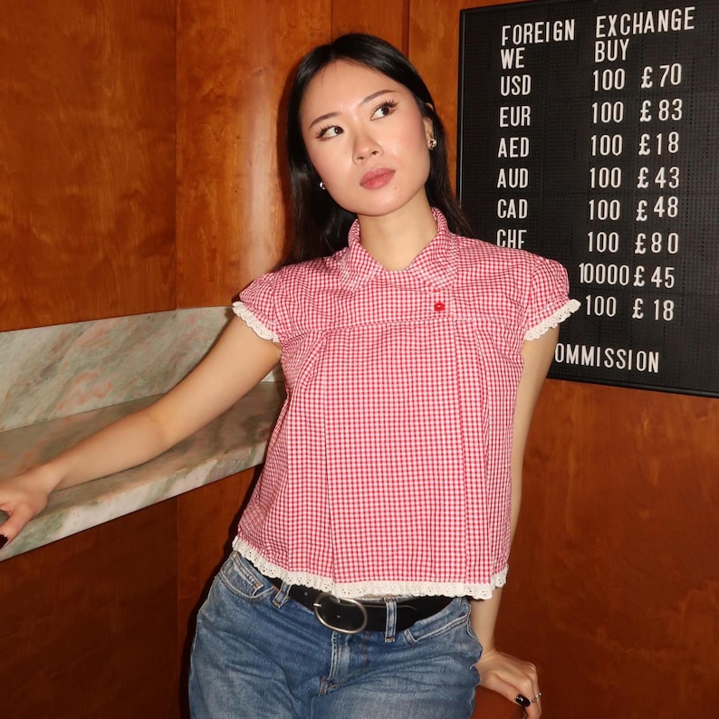 May include: A red and white gingham top with a white lace trim, paired with blue jeans and a black belt. The top has a small red button and a collared neckline. A sign in the background displays foreign exchange rates.