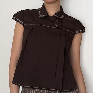 May include: A dark brown short-sleeved top with a collared neckline and two brown buttons. The top features white stitching along the collar, sleeves, and seams. The fabric is pleated on the front, creating a tailored look.