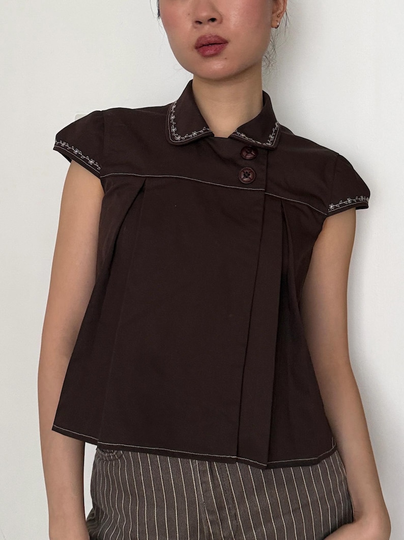 May include: A dark brown short-sleeved top with a collared neckline and two brown buttons. The top features white stitching along the collar, sleeves, and seams. The fabric is pleated on the front, creating a tailored look.