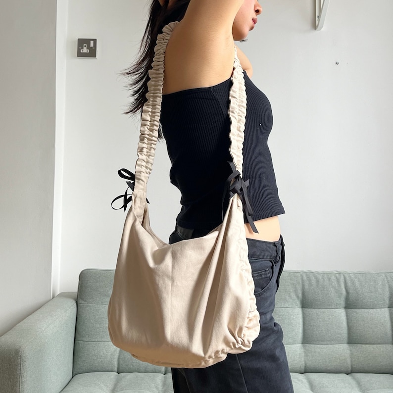 May include: A beige canvas shoulder bag with black ribbon ties and a gathered ruffle detail on the strap.