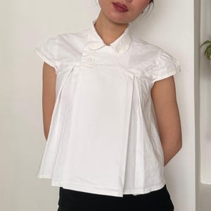 May include: White short-sleeved top with a unique asymmetrical design. The top features a button closure and a pleated front, creating a structured silhouette. The collar has a delicate embroidered detail. The garment is made of a crisp, white fabric.