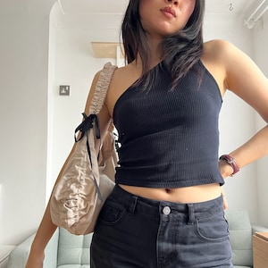 May include: A woman wearing a black one-shoulder top and black jeans. She is carrying a light beige fabric bag with a black ribbon tied around the handle.