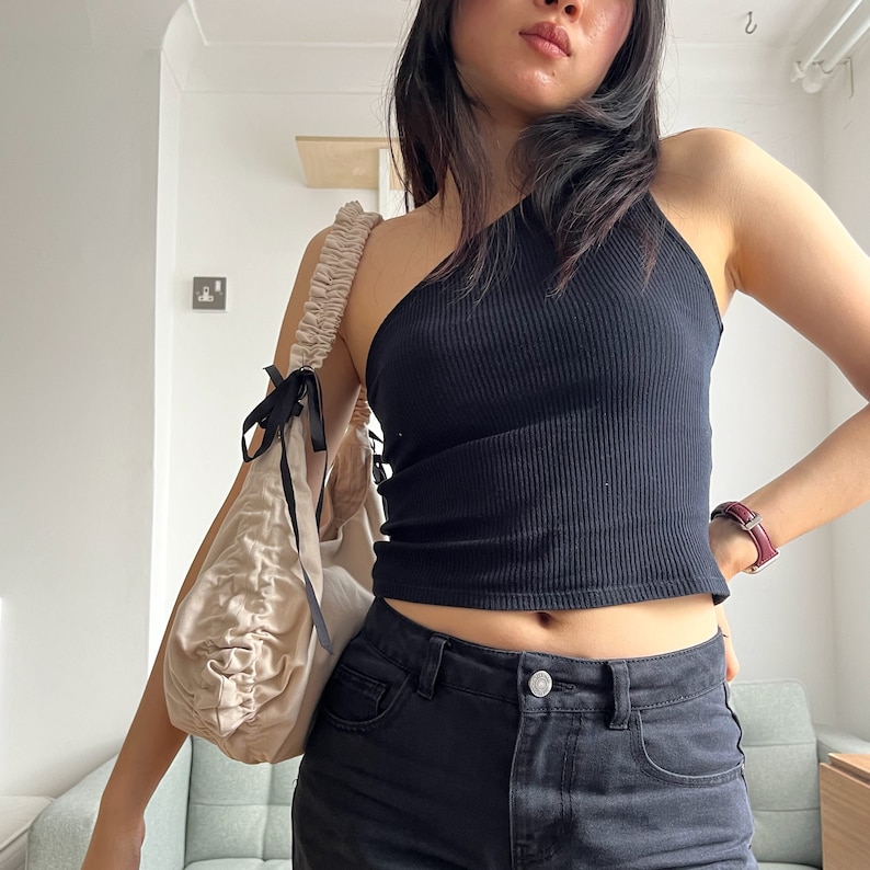 May include: A woman wearing a black one-shoulder top and black jeans. She is carrying a light beige fabric bag with a black ribbon tied around the handle.