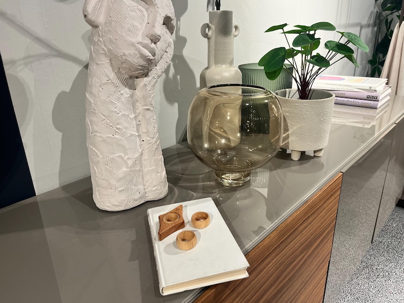 May include: A white ceramic sculpture of a face, a clear glass vase with a brown tint, a white ceramic planter with a green plant, and a white book with three wooden rings on top.