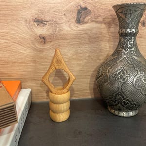 May include: A wooden ring holder with a unique design. The holder is made of light brown wood and has a ring-shaped opening in the center. The holder is sitting on a dark surface.