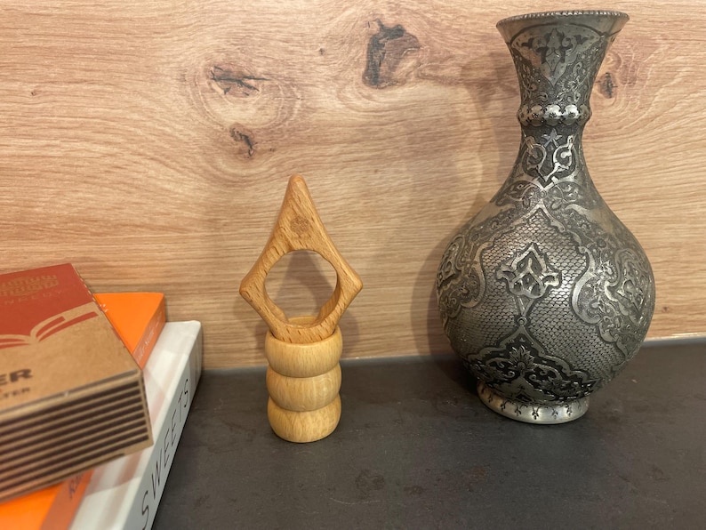 May include: A wooden ring holder with a unique design. The holder is made of light brown wood and has a ring-shaped opening in the center. The holder is sitting on a dark surface.