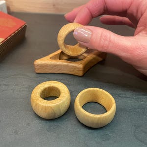 May include: Three wooden rings, two of which are lying on a dark surface, and one is being held by a hand. The ring being held is being placed on a wooden stand with a cutout in the center.