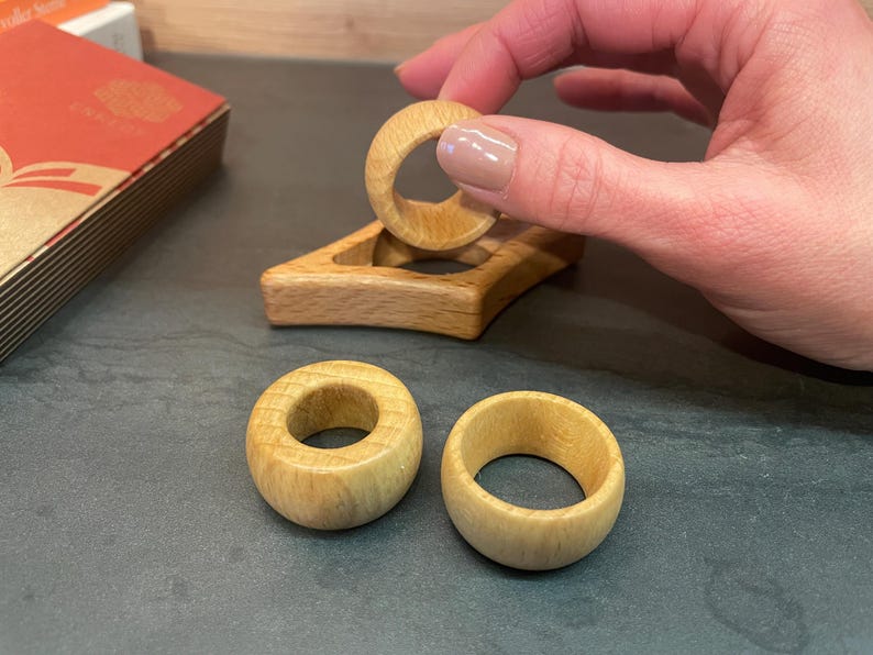 May include: Three wooden rings, two of which are lying on a dark surface, and one is being held by a hand. The ring being held is being placed on a wooden stand with a cutout in the center.