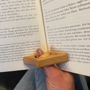 May include: A wooden thumb reading aid, also known as a page holder, is shown in use. The page holder is made of light brown wood and has a triangular shape. The page holder is being used to hold open a book with German text.
