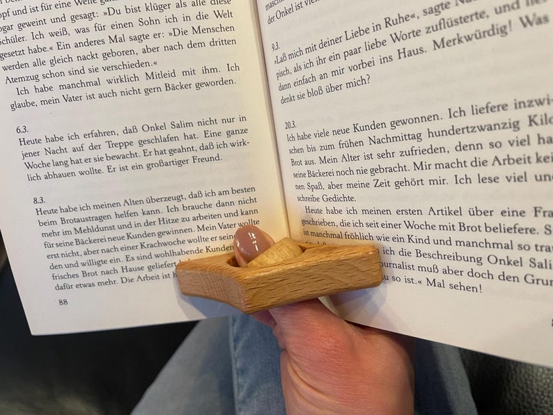 May include: A wooden thumb reading aid, also known as a page holder, is shown in use. The page holder is made of light brown wood and has a triangular shape. The page holder is being used to hold open a book with German text.