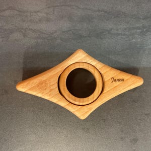 May include: A wooden bookmark shaped like a diamond with a round cutout in the center. The bookmark is engraved with the name "Janna".
