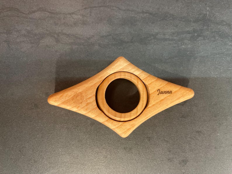 May include: A wooden bookmark shaped like a diamond with a round cutout in the center. The bookmark is engraved with the name "Janna".