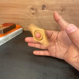 May include: A wooden fidget spinner with a round center and three blades. The spinner is being held by a hand.