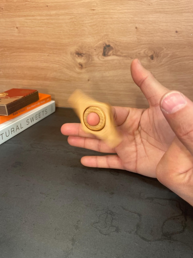 May include: A wooden fidget spinner with a round center and three blades. The spinner is being held by a hand.
