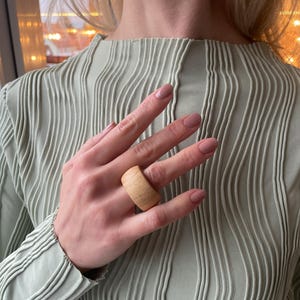 May include: A close-up of a hand wearing a large, light brown wooden ring. The ring is wide and has a smooth, natural finish.