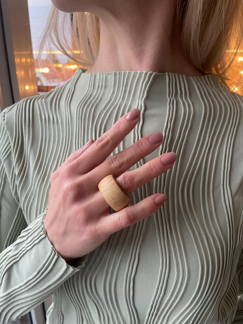 May include: A close-up of a hand wearing a large, light brown wooden ring. The ring is wide and has a smooth, natural finish.