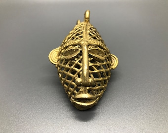 Puede incluir: Una máscara de metal de color dorado con un diseño geométrico. La máscara tiene una cara humana con un patrón de rejilla sobre los ojos y la nariz.