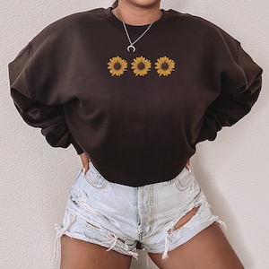 May include: A black cropped sweatshirt with three yellow sunflowers printed on the front. The sweatshirt is being worn with light blue denim shorts.