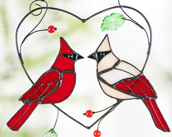 May include: A stained glass heart-shaped suncatcher with two cardinals perched on branches. The male cardinal is red and the female cardinal is white with red accents. The cardinals are facing each other and there are red berries on the branches.