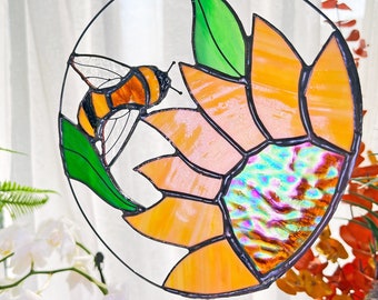 May include: A stained glass suncatcher featuring a stylized orange sunflower with a bee flying towards it. The sunflower has a green leaf and a iridescent center.